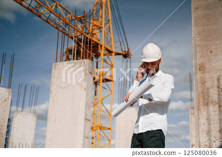 Talking by phone and holding document in hands. Man in uniform is working on the construction site 125080036