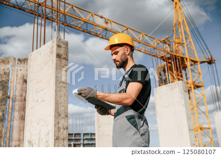 Standing against yellow crane and holding notepad with documents. Man in uniform is working on the construction site Standing against yellow crane and holding notepad with documents. Man in uniform is working on the construction site 125080107