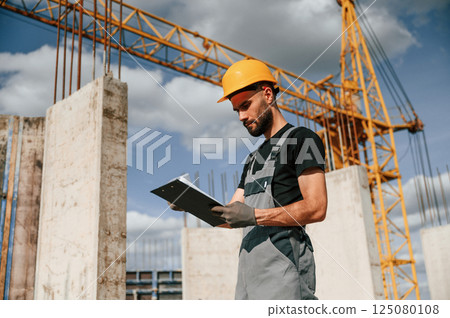 Standing against yellow crane and holding notepad with documents. Man in uniform is working on the construction site Standing against yellow crane and holding notepad with documents. Man in uniform is working on the construction site 125080108