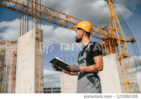 Standing against yellow crane and holding notepad with documents. Man in uniform is working on the construction site Standing against yellow crane and holding notepad with documents. Man in uniform is working on the construction site 125080109
