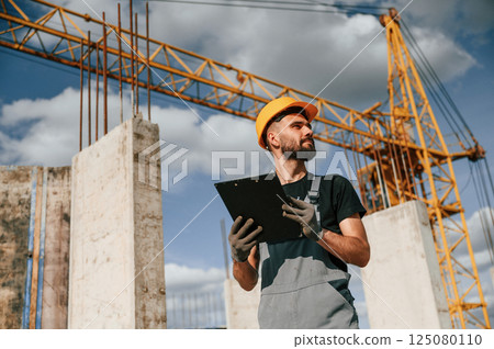 Standing against yellow crane and holding notepad with documents. Man in uniform is working on the construction site Standing against yellow crane and holding notepad with documents. Man in uniform is working on the construction site 125080110