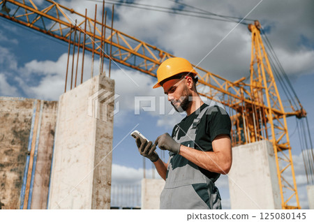 With phone in hands. Standing against crane. Man in uniform is working on the construction site 125080145
