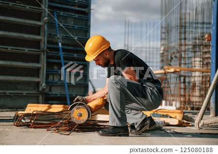 Sitting and cutting metal pieces by grinding machine. Man in uniform is working on the construction site Sitting and cutting metal pieces by grinding machine. Man in uniform is working on the construction site 125080188