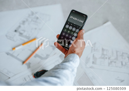 Calculating numbers by using smartphone. Plan is on the floor. The man is making repairs in the apartment Calculating numbers by using smartphone. Plan is on the floor. The man is making repairs in the apartment 125080309
