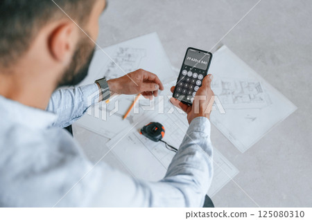 Calculating numbers by using smartphone. Plan is on the floor. The man is making repairs in the apartment 125080310