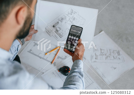 Calculating numbers by using smartphone. Plan is on the floor. The man is making repairs in the apartment Calculating numbers by using smartphone. Plan is on the floor. The man is making repairs in the apartment 125080312