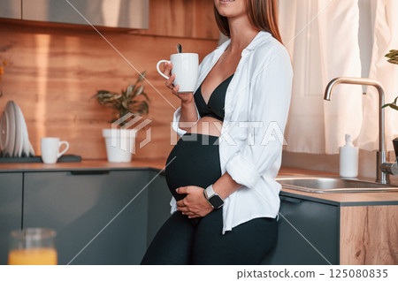 Standing on the kitchen with cup of drink. Beautiful pregnant woman is indoors at home Standing on the kitchen with cup of drink. Beautiful pregnant woman is indoors at home 125080835