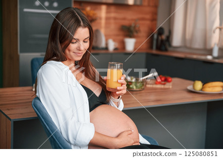 Touching belly and holding glass with orange juice. Beautiful pregnant woman is indoors at home Touching belly and holding glass with orange juice. Beautiful pregnant woman is indoors at home 125080851