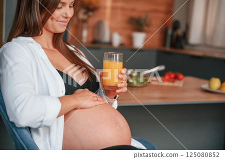 Touching belly and holding glass with orange juice. Beautiful pregnant woman is indoors at home 125080852