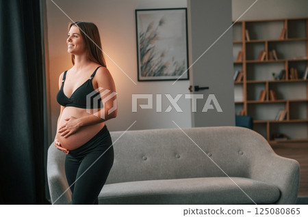 Standing in the domestic room and touching the belly. Beautiful pregnant woman is indoors at home 125080865