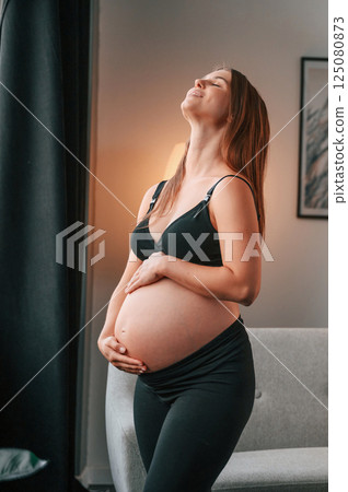 Standing and having positive mood. Beautiful pregnant woman is indoors at home Standing and having positive mood. Beautiful pregnant woman is indoors at home 125080873