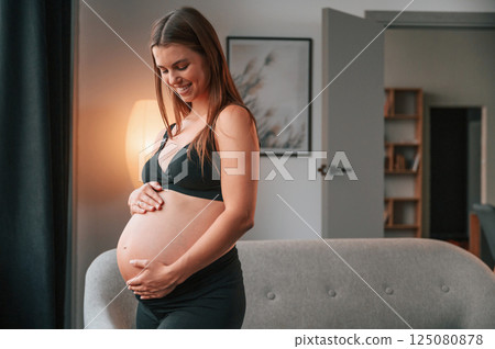 Standing near the sofa and smiling. Beautiful pregnant woman is indoors at home Standing near the sofa and smiling. Beautiful pregnant woman is indoors at home 125080878