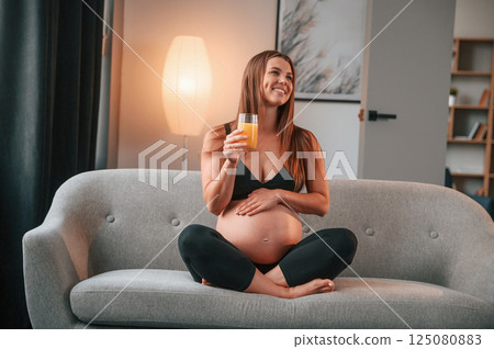 Sitting on the sofa and drinking orange juice. Beautiful pregnant woman is indoors at home 125080883