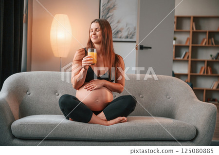 With orange juice in hand. On the sofa. Beautiful pregnant woman is indoors at home With orange juice in hand. On the sofa. Beautiful pregnant woman is indoors at home 125080885