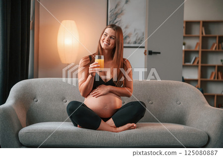 With orange juice in hand. On the sofa. Beautiful pregnant woman is indoors at home With orange juice in hand. On the sofa. Beautiful pregnant woman is indoors at home 125080887