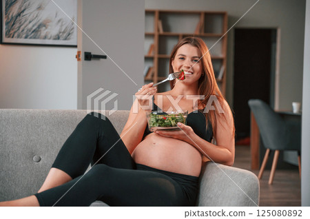 Sitting on the sofa and eating salad. Beautiful pregnant woman is indoors at home Sitting on the sofa and eating salad. Beautiful pregnant woman is indoors at home 125080892