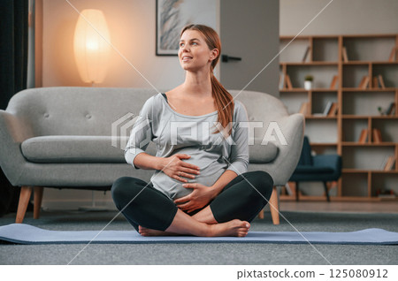 Sitting on the yoga mat. Beautiful pregnant woman is indoors at home 125080912