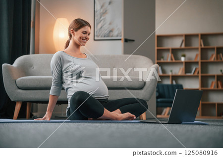 Watching yoga lessons by using laptop. Beautiful pregnant woman is indoors at home 125080916