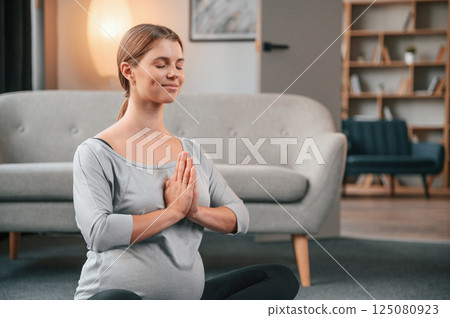 Sitting and doing meditation. Beautiful pregnant woman is indoors at home 125080923