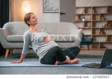 On yoga mat with laptop. Beautiful pregnant woman is indoors at home 125080927
