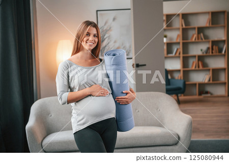 With yoga mat in hand. Beautiful pregnant woman is indoors at home 125080944