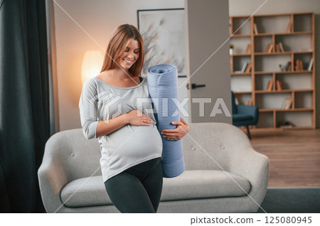 With yoga mat in hand. Beautiful pregnant woman is indoors at home 125080945