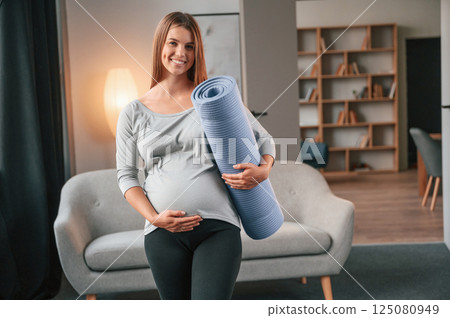Blue colored yoga mat is in hand. Beautiful pregnant woman is indoors at home 125080949