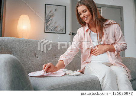 With little clothes for a baby on the sofa. Beautiful pregnant woman is indoors at home 125080977