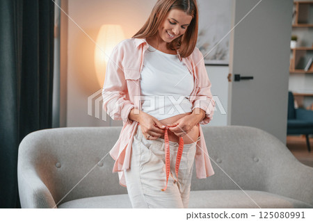 With measuring tape. Around the waist. Beautiful pregnant woman is indoors at home 125080991