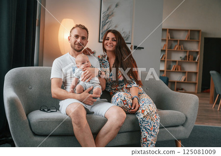Sitting on the sofa. Happy couple with their newborn boy is at home together Sitting on the sofa. Happy couple with their newborn boy is at home together 125081009