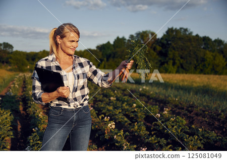 Document with information and carrots in hand. Woman is on the agricultural field at daytime 125081049
