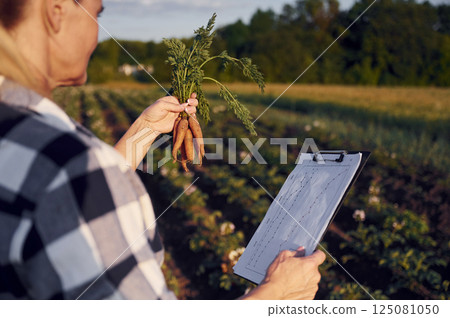 Document with information and carrots in hand. Woman is on the agricultural field at daytime 125081050