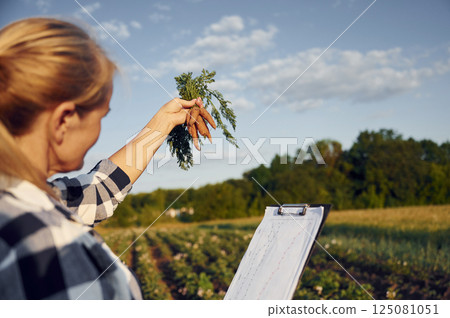 Document with information and carrots in hand. Woman is on the agricultural field at daytime 125081051