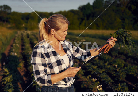 Standing with notepad and holding carrots. Woman is on the agricultural field at daytime 125081272