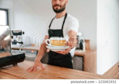 Standing and holding cup. Cafe worker in white shirt and black apron is indoors 125081932