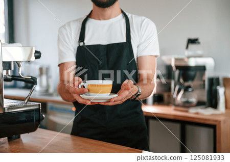 Cup is in the hands. Cafe worker in white shirt and black apron is indoors 125081933
