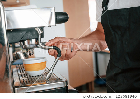 Using coffee machine. Cafe worker in white shirt and black apron is indoors 125081940