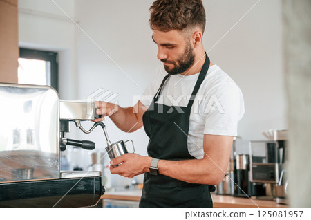 Standing and pouring coffee. Cafe worker in white shirt and black apron is indoors Standing and pouring coffee. Cafe worker in white shirt and black apron is indoors 125081957