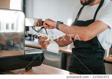 Cleaning coffee machine. Cafe worker in white shirt and black apron is indoors 125081972
