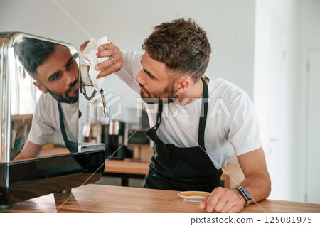 Cleaning coffee machine. Cafe worker in white shirt and black apron is indoors 125081975