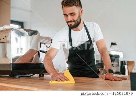 Cleaning table. Cafe worker in white shirt and black apron is indoors Cleaning table. Cafe worker in white shirt and black apron is indoors 125081979