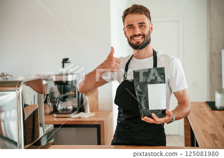 Holding package of raw coffee and showing thumb up. Cafe worker in white shirt and black apron is indoors Holding package of raw coffee and showing thumb up. Cafe worker in white shirt and black apron is indoors 125081980