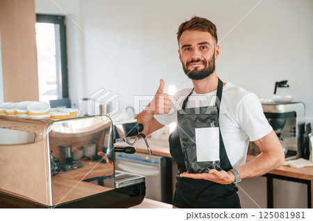 Holding package of raw coffee and showing thumb up. Cafe worker in white shirt and black apron is indoors 125081981