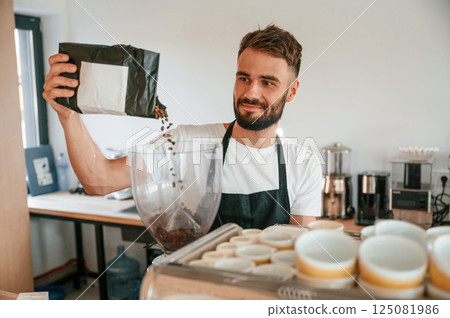 Pours whole coffee beans. Cafe worker in white shirt and black apron is indoors 125081986