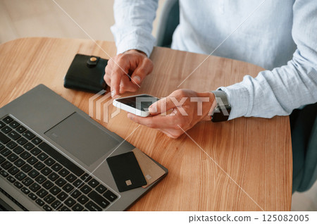 Credit card is on the laptop. Close up view of man that sitting by the table and working by using smartphone 125082005