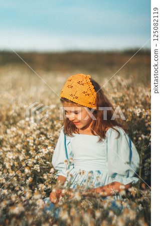 Smiling girl in blue linen dress and natural yellow knitted handmade kerchief in daisy field in summer day Smiling girl in blue linen dress and natural yellow knitted handmade kerchief in daisy field in summer day 125082119