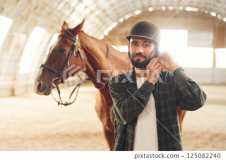 In protective hat. Jockey equipment. Young man with a horse is in the hangar In protective hat. Jockey equipment. Young man with a horse is in the hangar 125082240