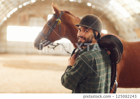 In protective hat. Jockey equipment. Young man with a horse is in the hangar 125082243