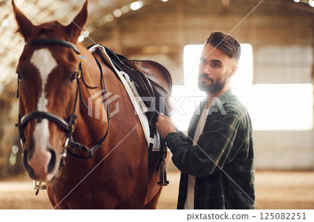Installing the saddle. Young man with a horse is in the hangar 125082251