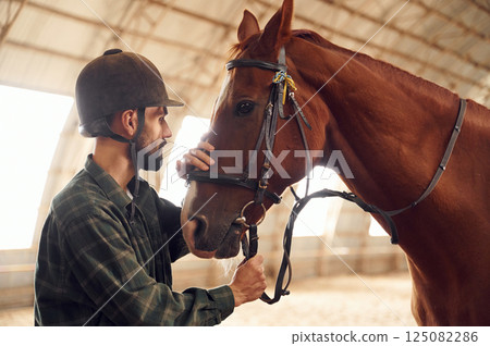 Standing and holding animal. Young man with a horse is in the hangar 125082286
